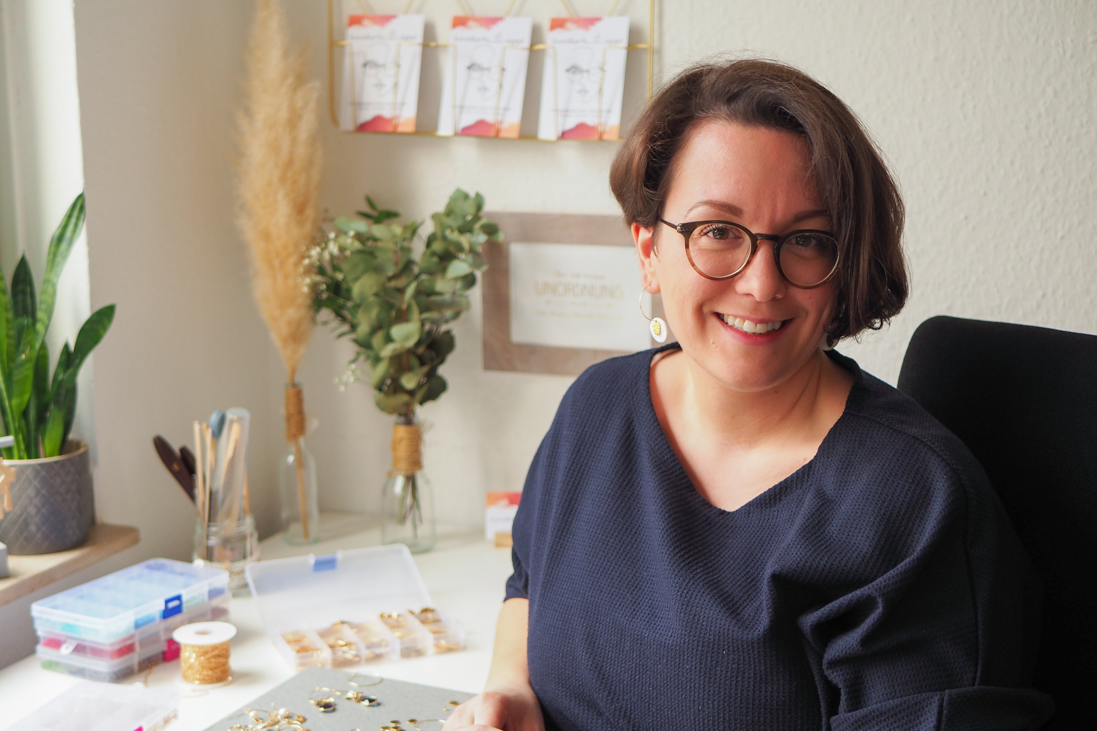 Eine Person mit kurzen Haaren und Brille sitzt lächelnd an einem Schreibtisch. Es ist Britta Albracht, die Gründerin von Kandierte Äpfel. Auf dem Tisch liegen Bastelmaterialien und im Hintergrund stehen Pflanzen. Die Umgebung ist hell und einladend.