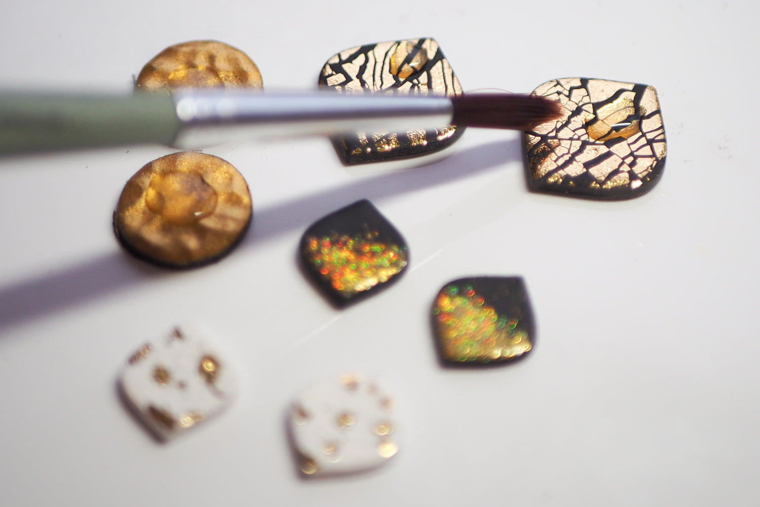 Mit einem Pinsel wird vorsichtig Lack auf mit Blattgold verzierte Polymerton Teile aufgetragen. Die Teile haben verschiedene  Formen und Farben, darunter Gold, Schwarz und Weiß.