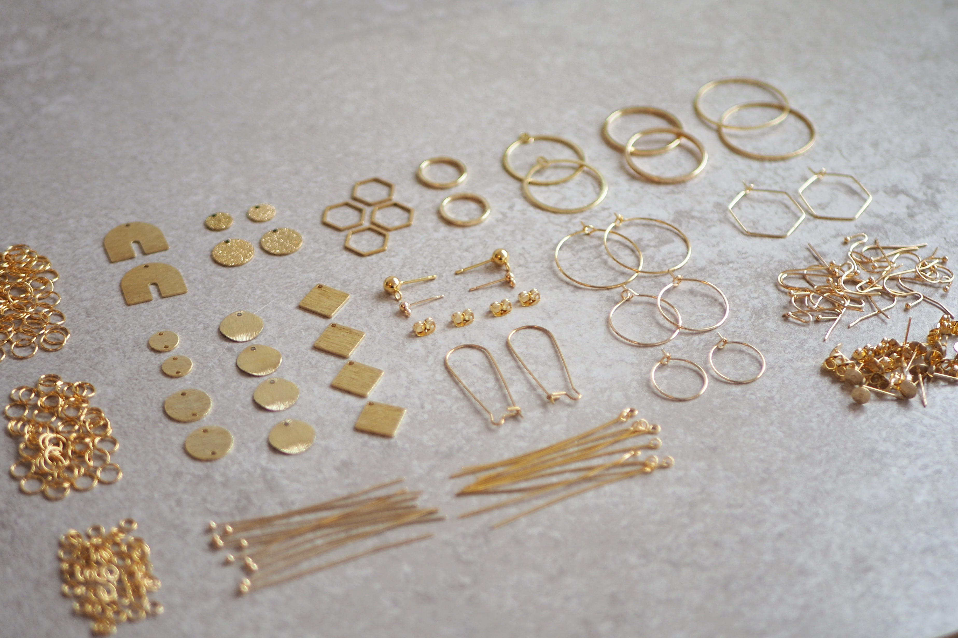 Eine Auswahl an Schmuckzubehör in Gold, darunter verschiedene Ohrringhaken, Creolen, geometrische Anhänger und Rahmen, die ordentlich auf einer hellgrauen Oberfläche präsentiert wurden. Sie können im Onlineshop Kandierte Äpfel gekauft werden.